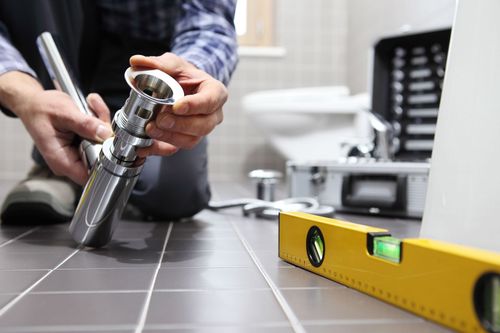 Hands installing a chrome pipe fitting on a tiled floor beside a yellow level tool