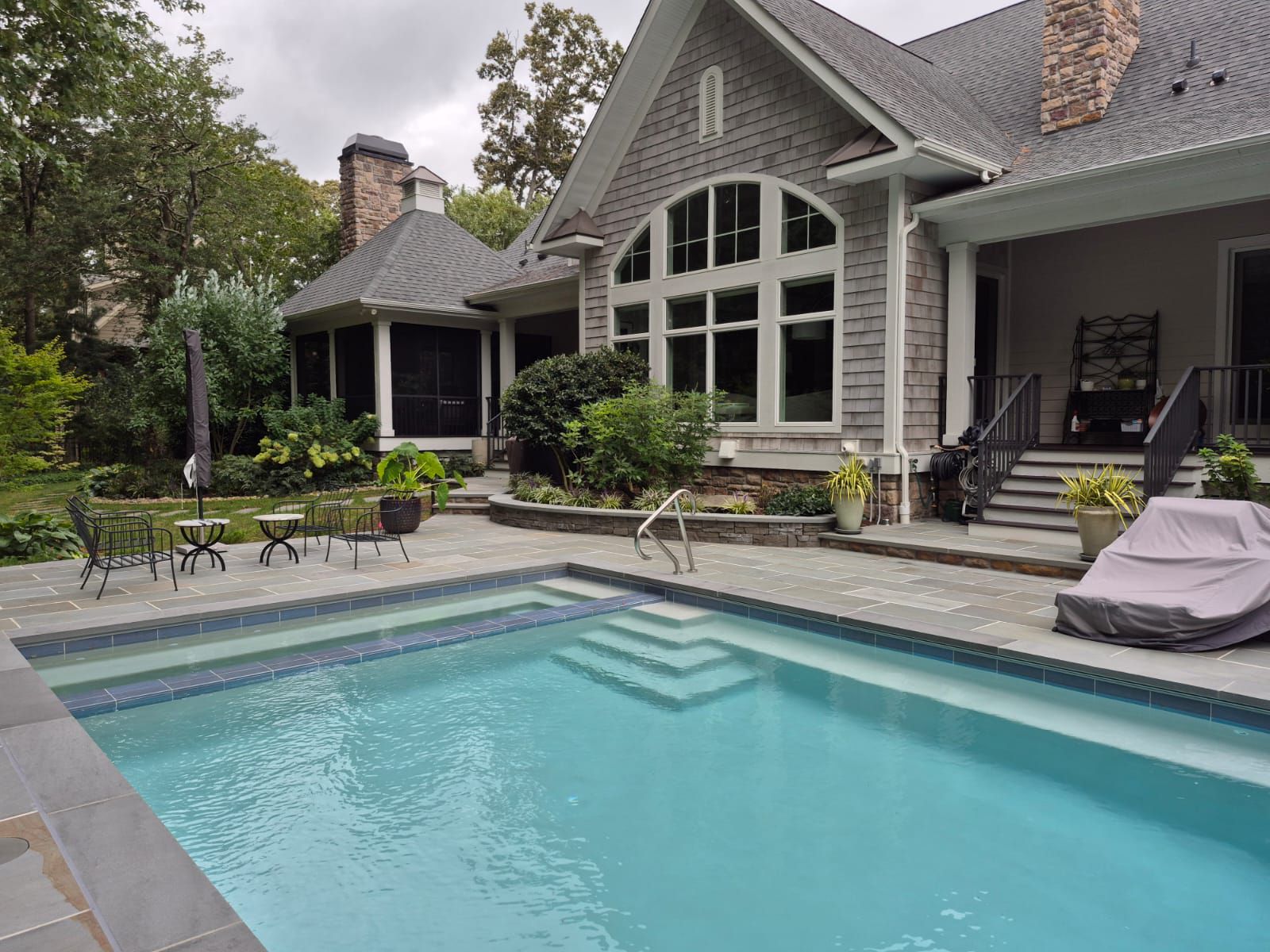 A swimming pool with steps in the foreground sits on a stone patio behind a large, stone-faced house with a arched window.