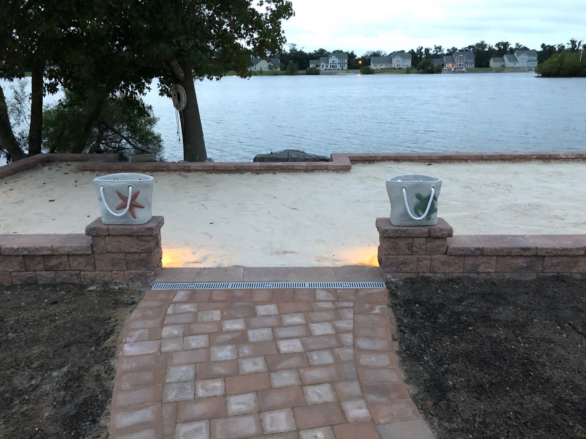 A brick walkway leads to a sandy beach with a lake in the background