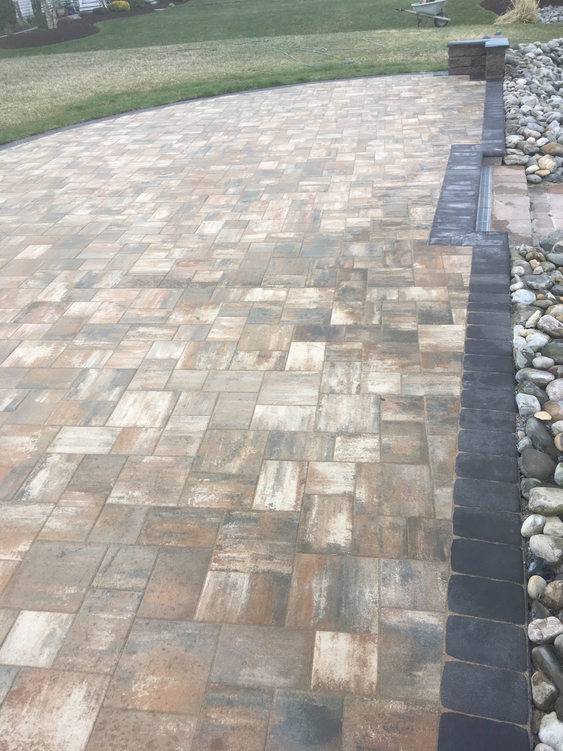 A brick driveway with a black border is surrounded by rocks and grass.