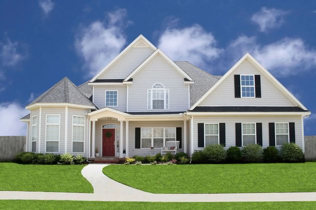 A two-story light gray suburban home with black shutters, a red front door, and a grassy front lawn under a blue sky.