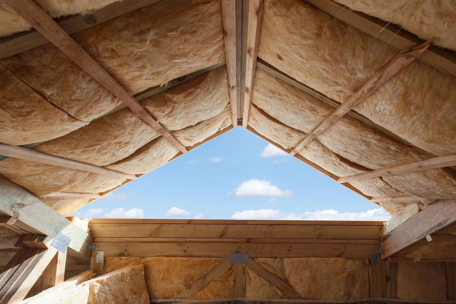 Attic insulation installed between wooden roof rafters, with an open triangular gap showing a blue sky and white clouds.