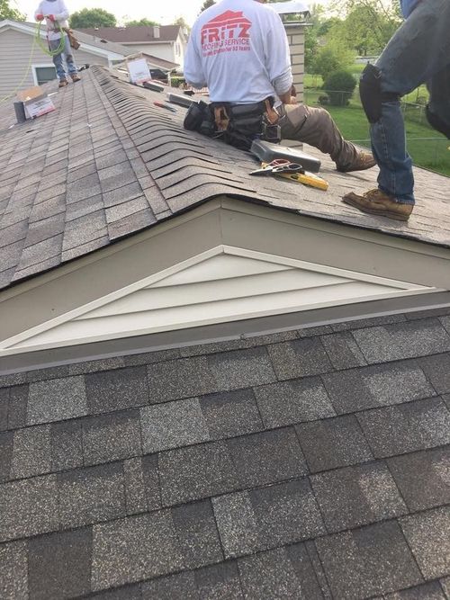 Two roofing contractors work on a shingled roof next to a triangular gable vent.