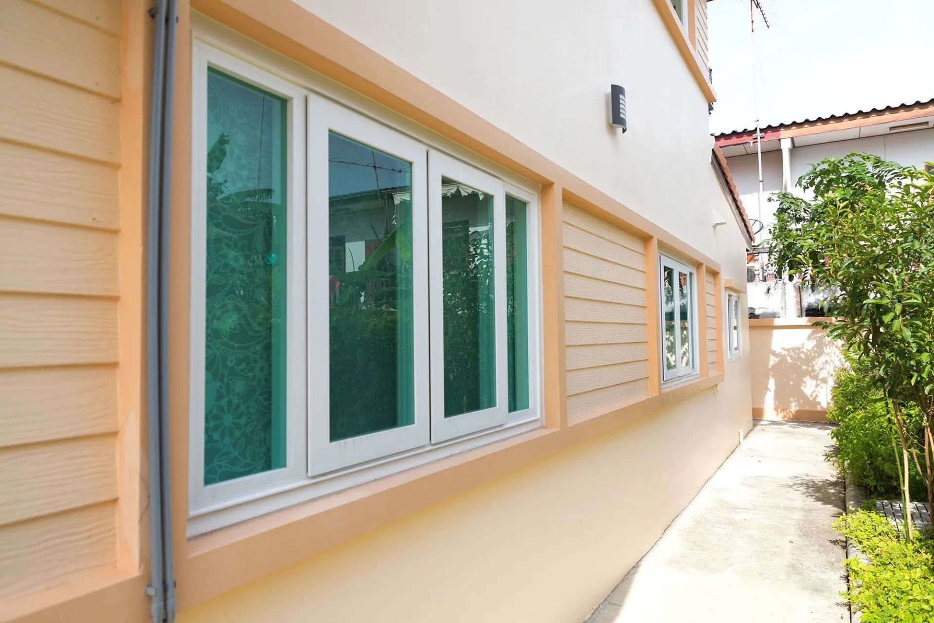 A side view of a cream-colored house with large white-framed windows, horizontal wall paneling, and a paved walkway.