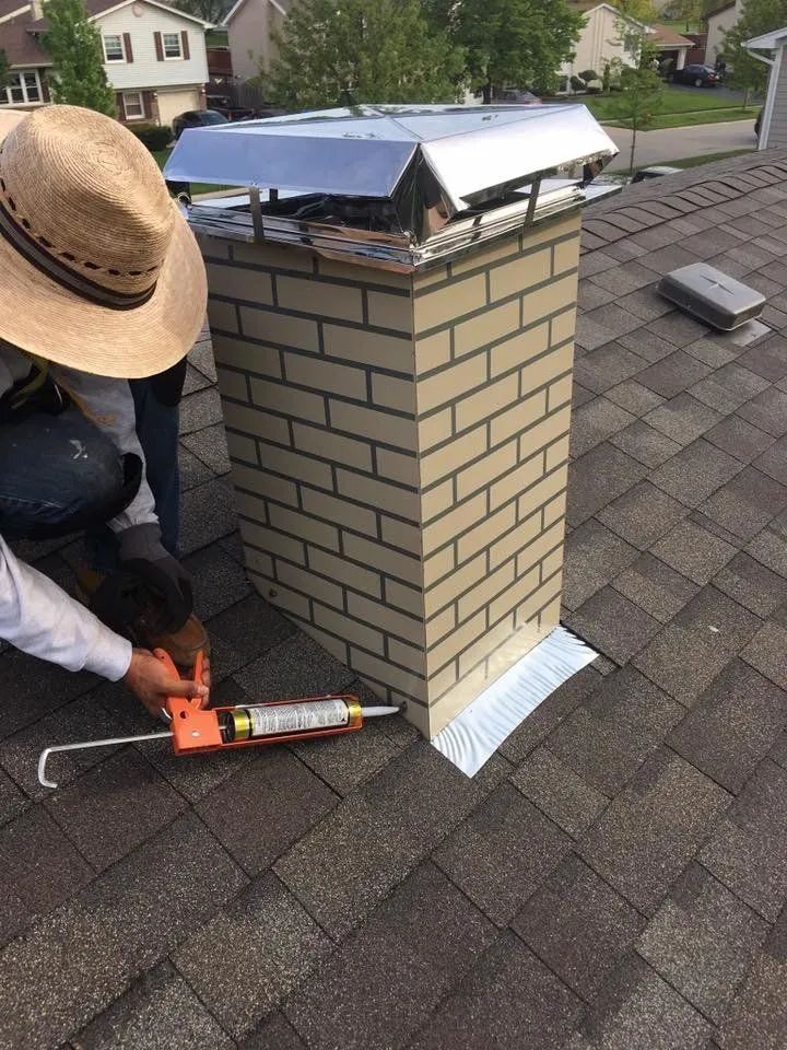 A person in a straw hat caulks the base of a brick chimney where it meets the asphalt shingle roof.