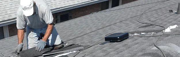 A worker in a white hat and gray shirt kneels on a shingled roof, repairing a section of roofing material.
