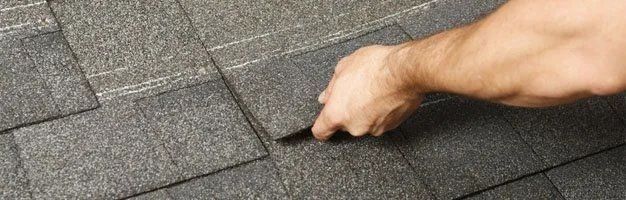 A hand inspects dark grey asphalt shingles installed on a roof.