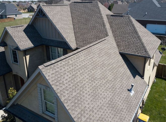 An aerial, high-angle view of a residential house with a complex, multi-gabled shingled roof and tan stucco siding.