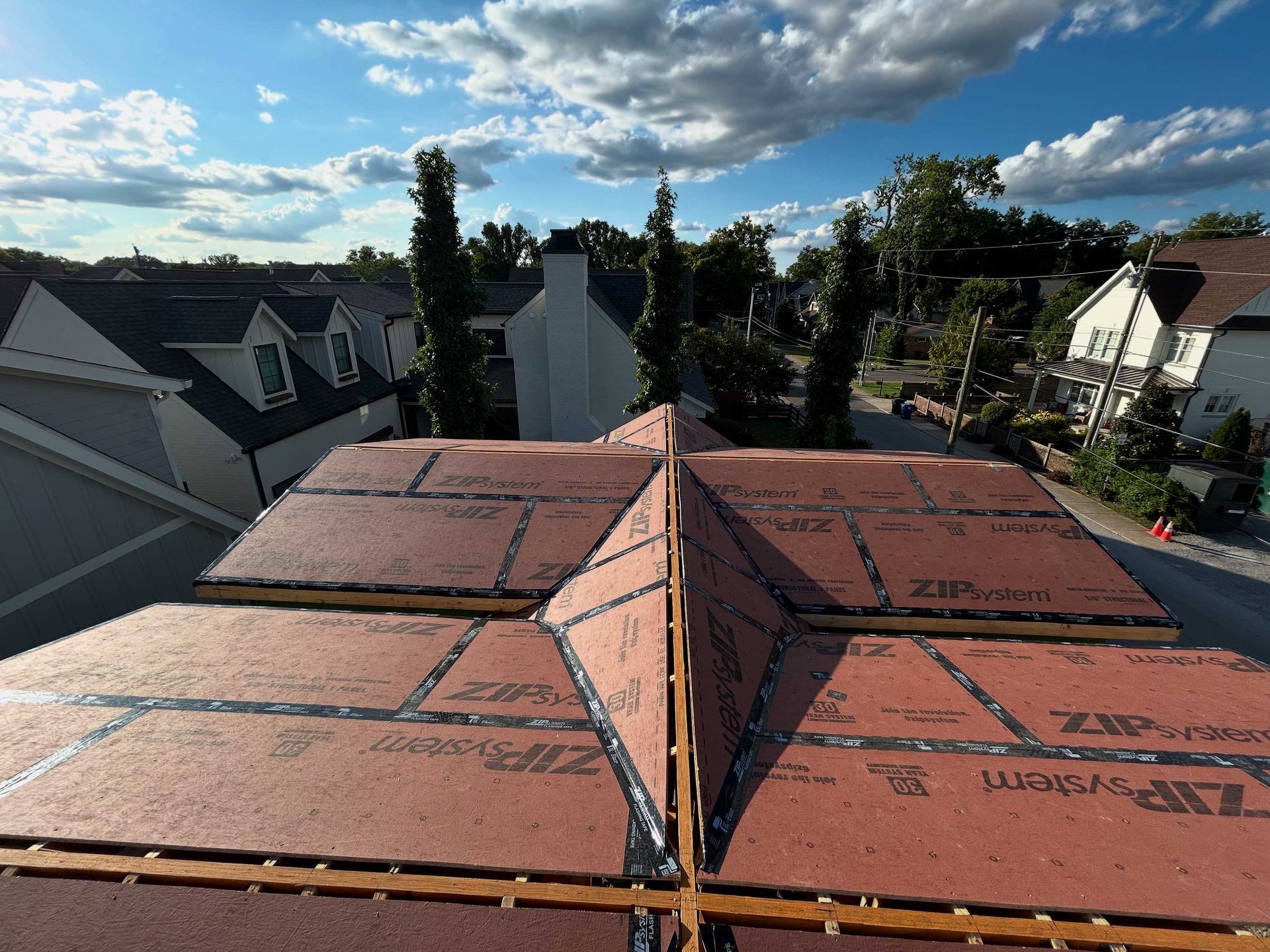 View of a roof with exposed red sheathing. Street and houses in the background under a blue sky with clouds.