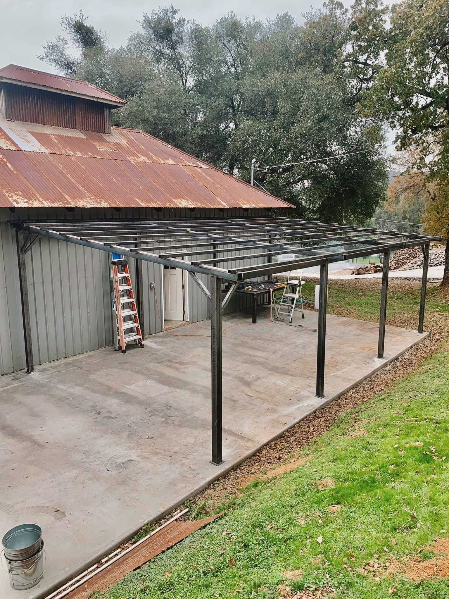 Pergola attached to a rustic building with a concrete patio; surrounded by grass and trees.