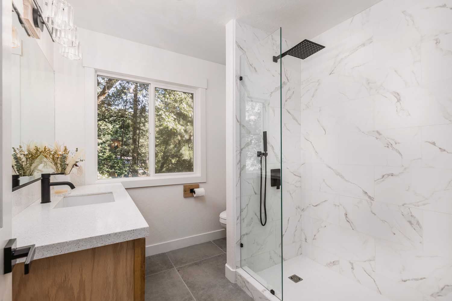 Modern bathroom with white marble shower, wood vanity, and window with trees.