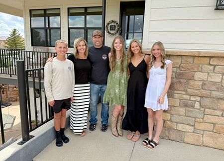 Family posing on porch: boy, woman in striped skirt, man, three girls, standing in front of a house.