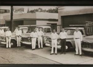 Men in white uniforms stand beside vintage trucks outside a building.