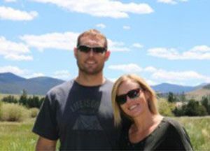 Couple wearing sunglasses smiles outdoors; mountains and blue sky in background.