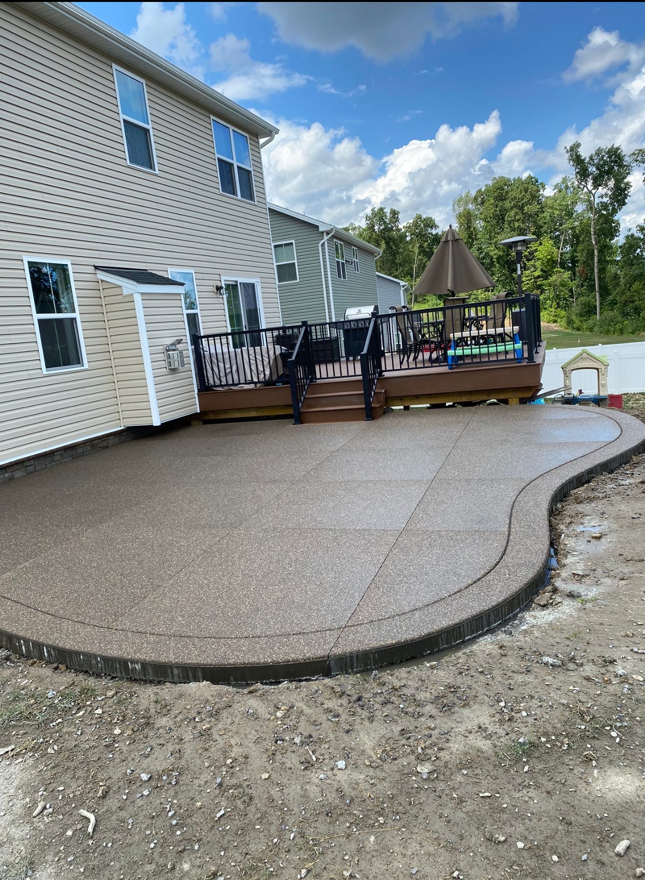 Backyard with a brown concrete patio, wooden deck, and a beige house on a sunny day.