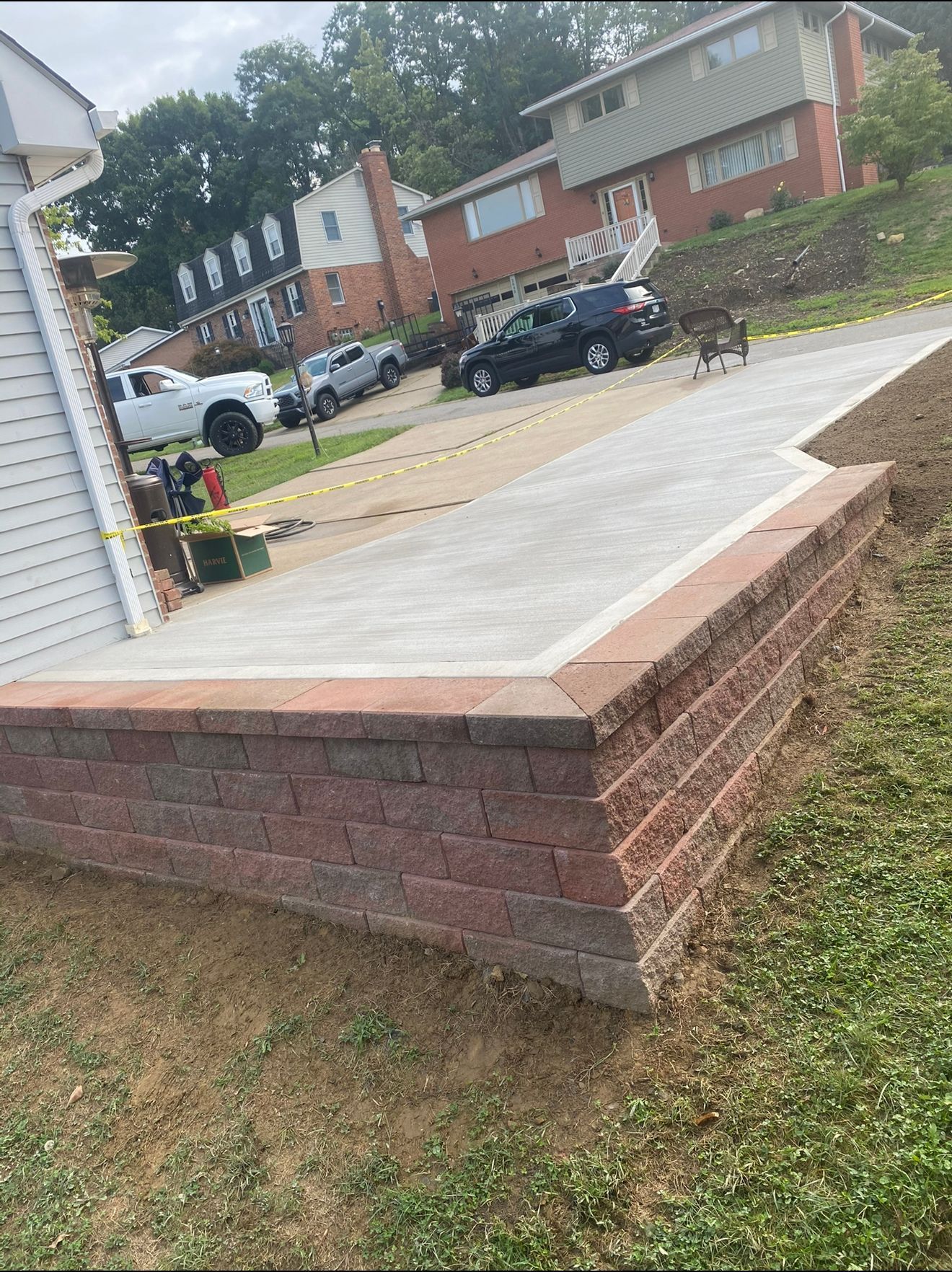 Brick retaining wall with concrete patio in front of houses; cars parked in background.