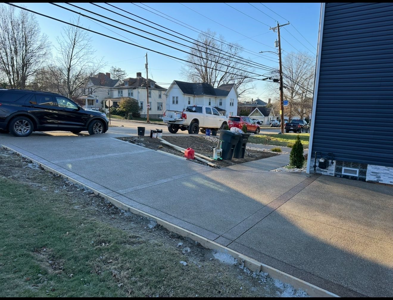 Concrete driveway with parked vehicles and a small garden bed beside a blue house on a residential street.