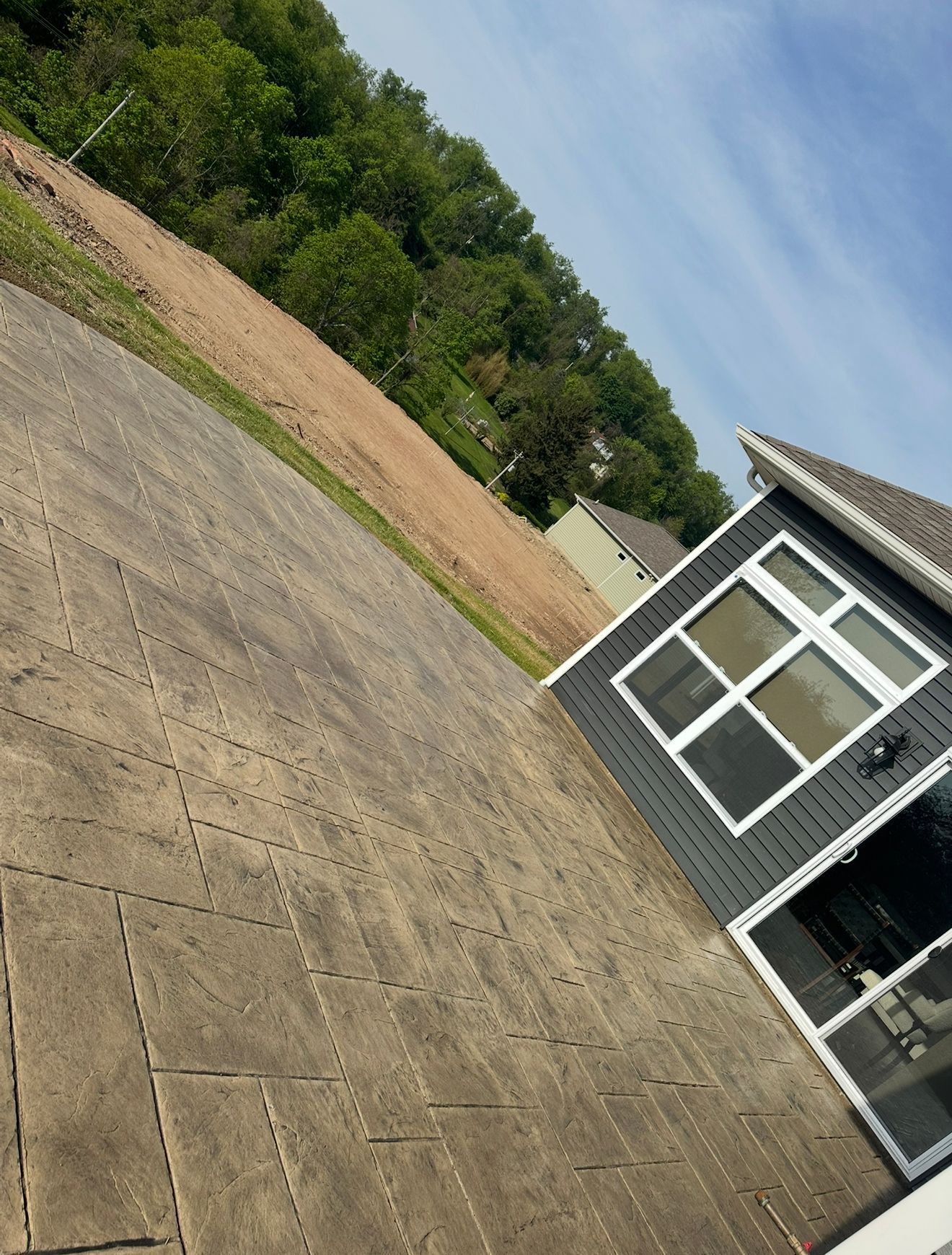Gray stamped concrete patio next to a house with a dark gray exterior, trees, and a blue sky.