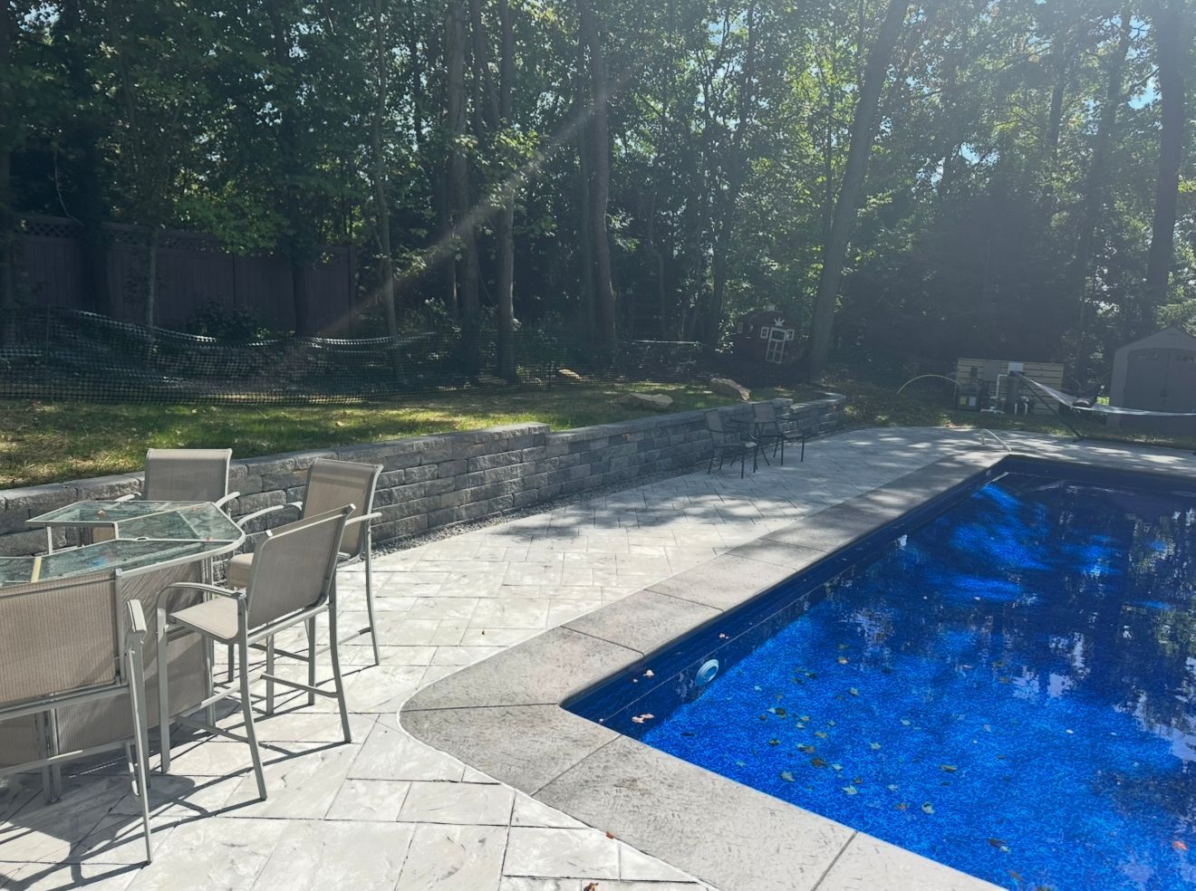 Poolside patio with table, chairs, and rectangular pool; trees in the background.