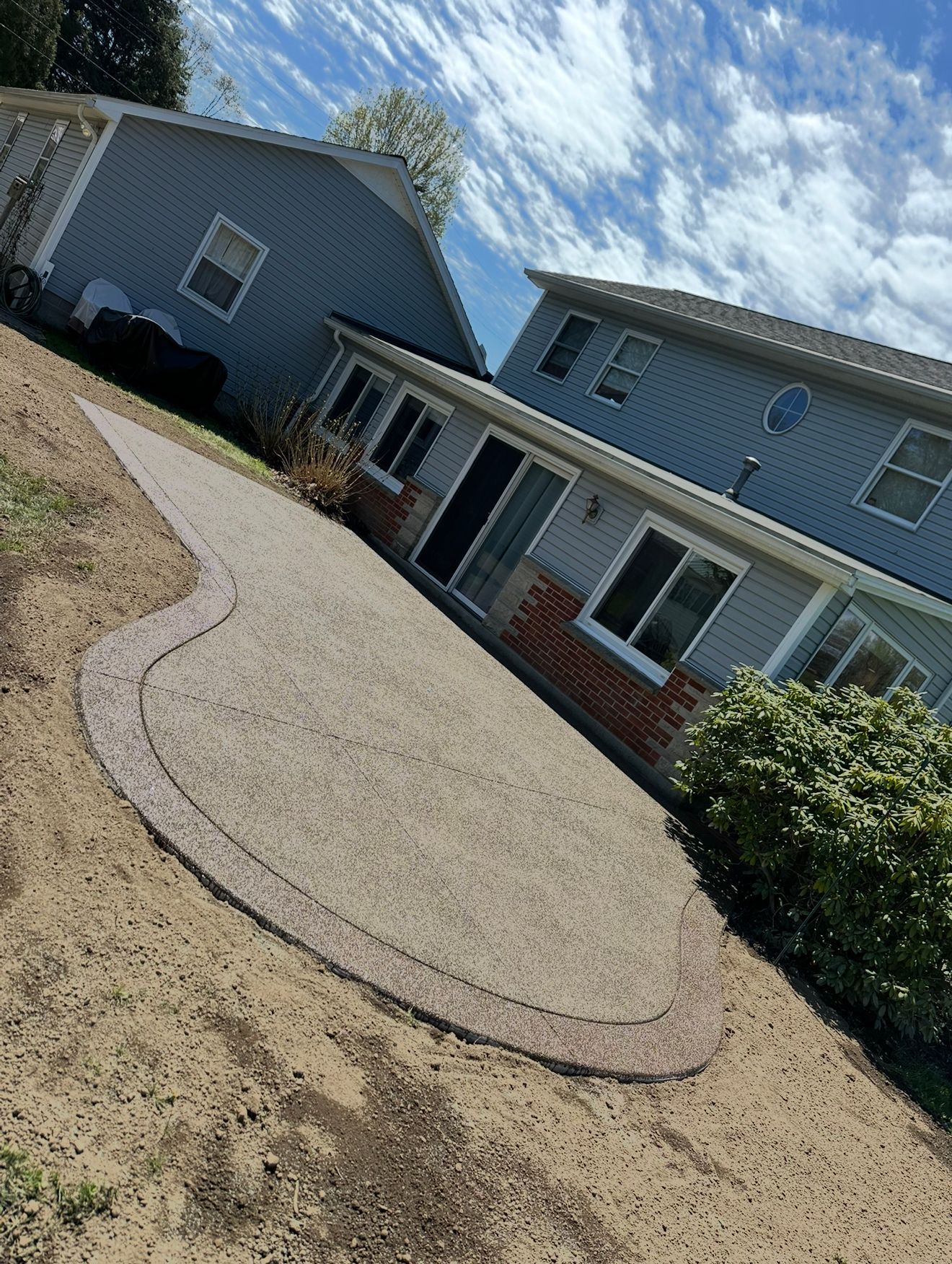 Backyard patio with light-colored concrete and decorative border, next to a two-story house under a blue sky.