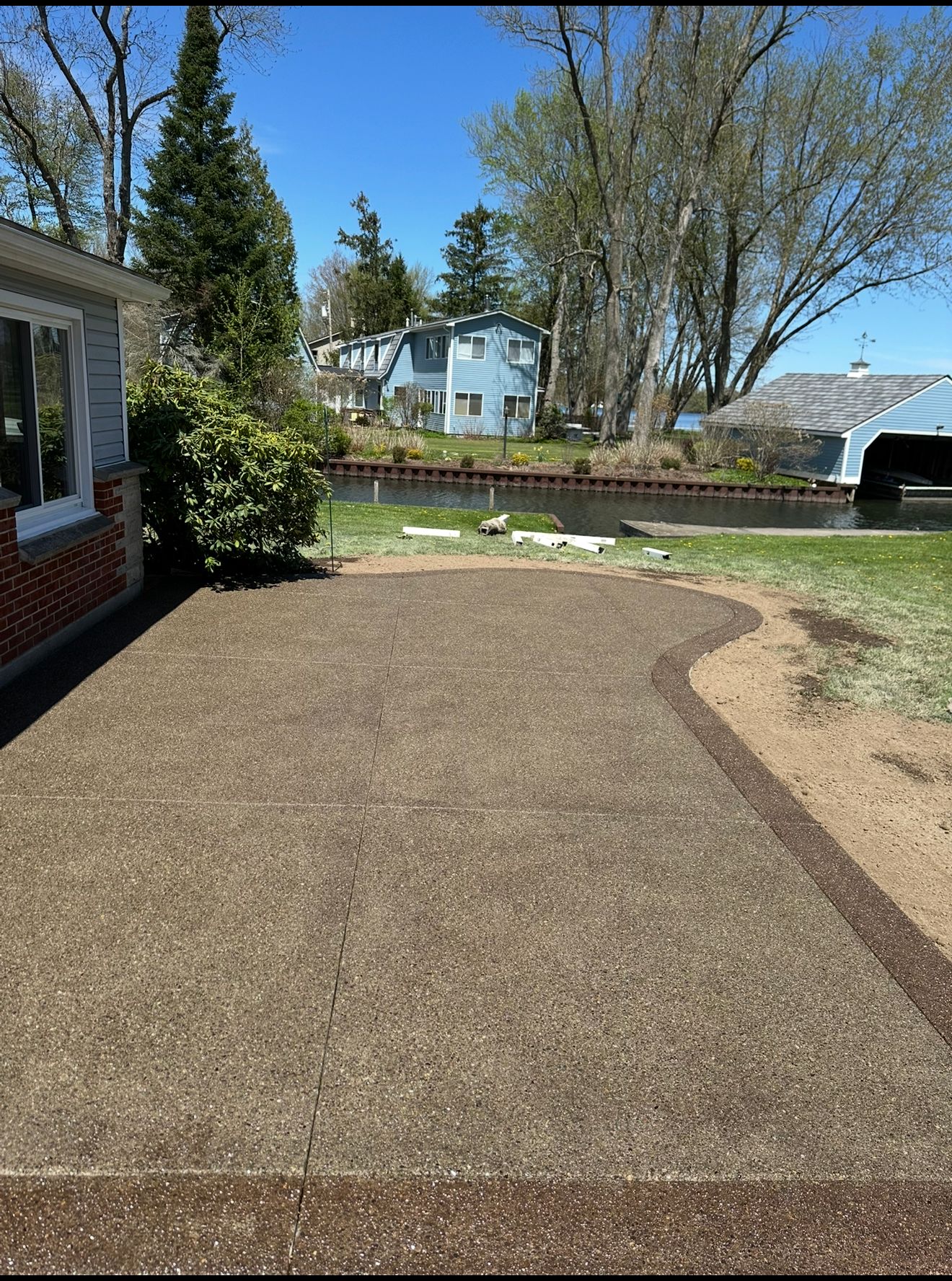 Patio with gravel surface next to a brick house and grassy area; blue sky and trees in the background.