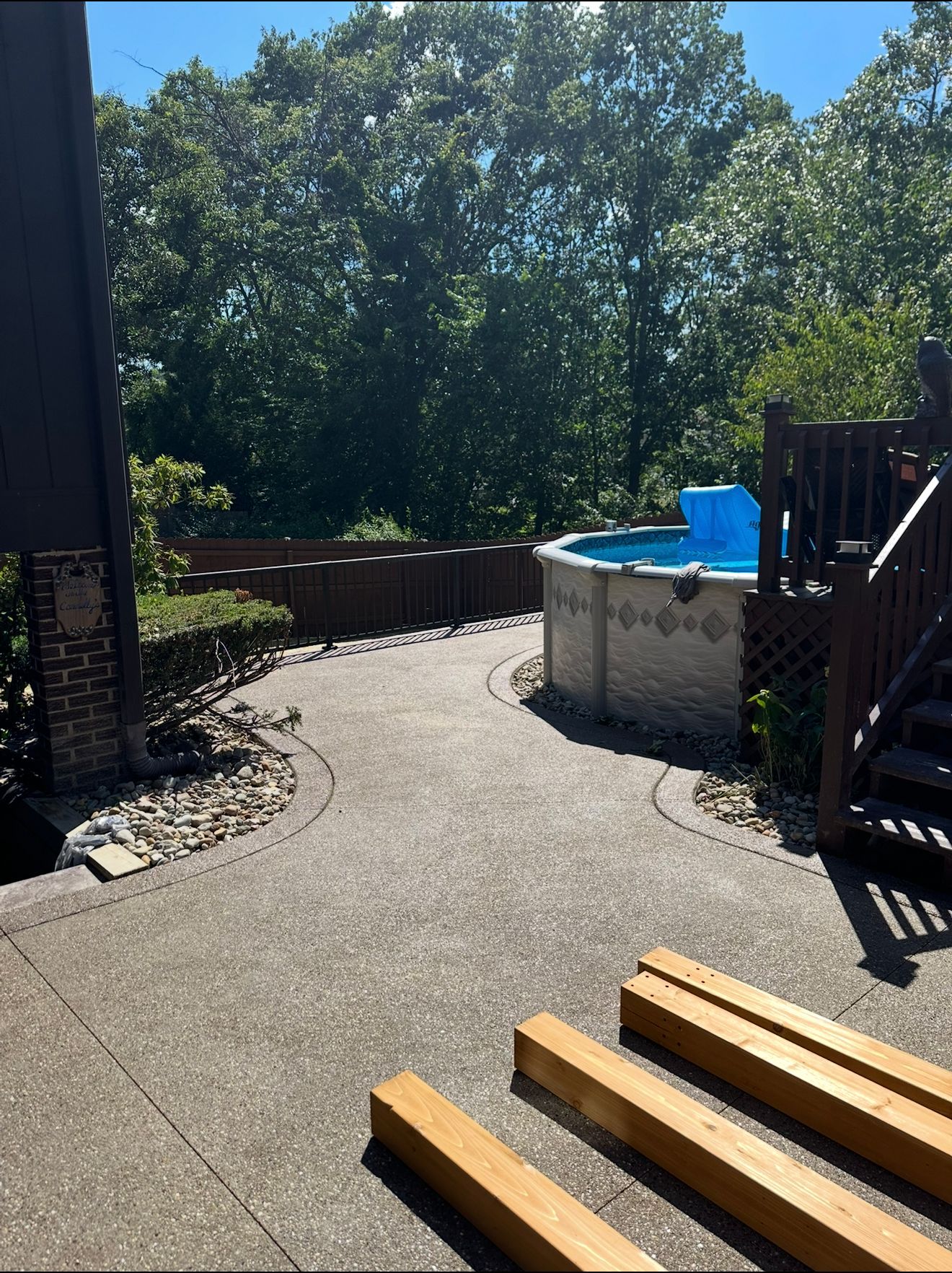 Concrete patio with a pool, fence, and trees in the background. Wooden beams in the foreground.