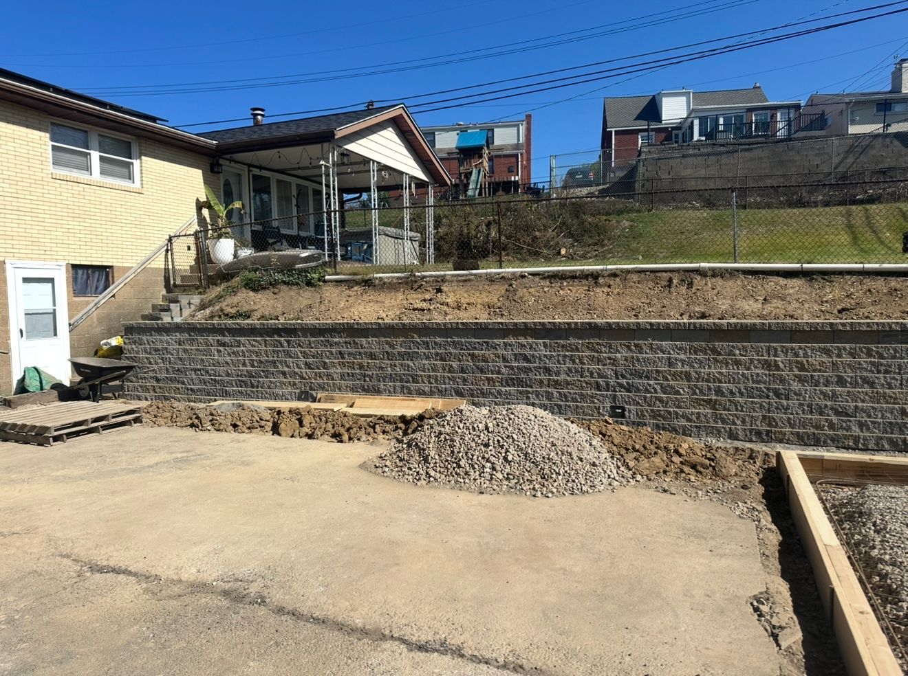 Retaining wall under construction with gravel pile, beside a light-colored building and hillside in a sunny environment.