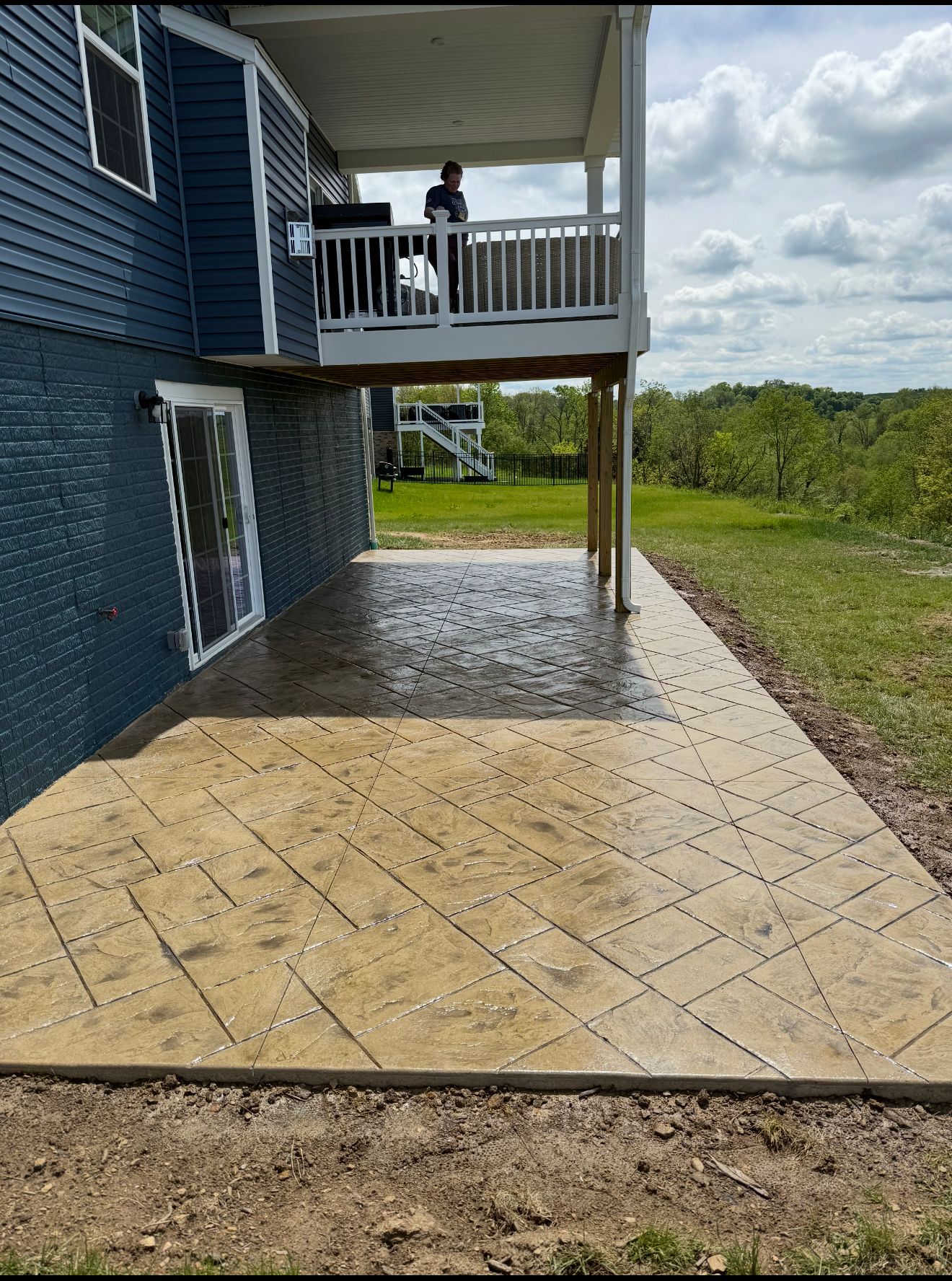 A concrete patio with a patterned design adjoins a two-story home with a blue exterior and a deck.