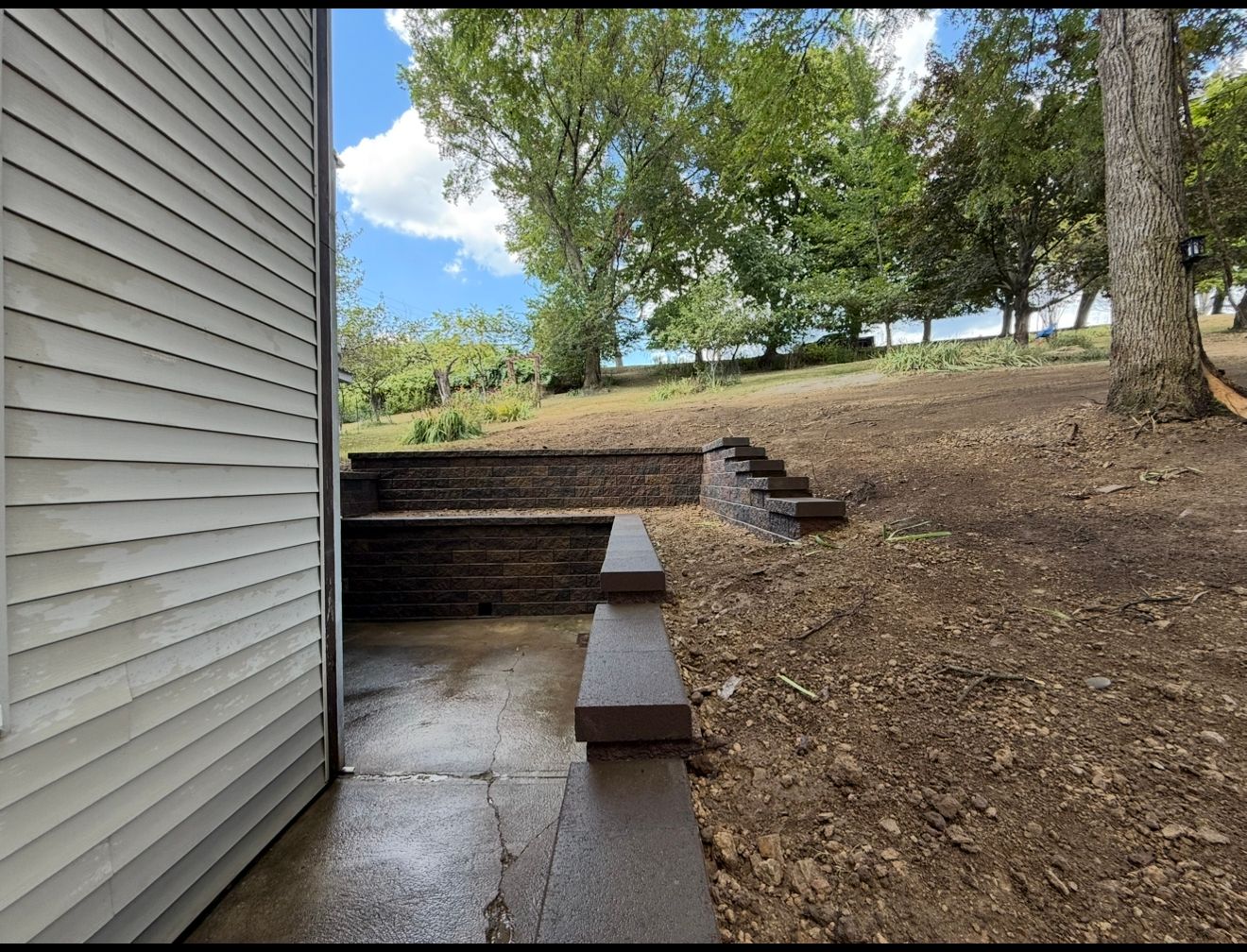 Exterior view of a house with a concrete patio, steps, and retaining wall. Trees and sky in the background.