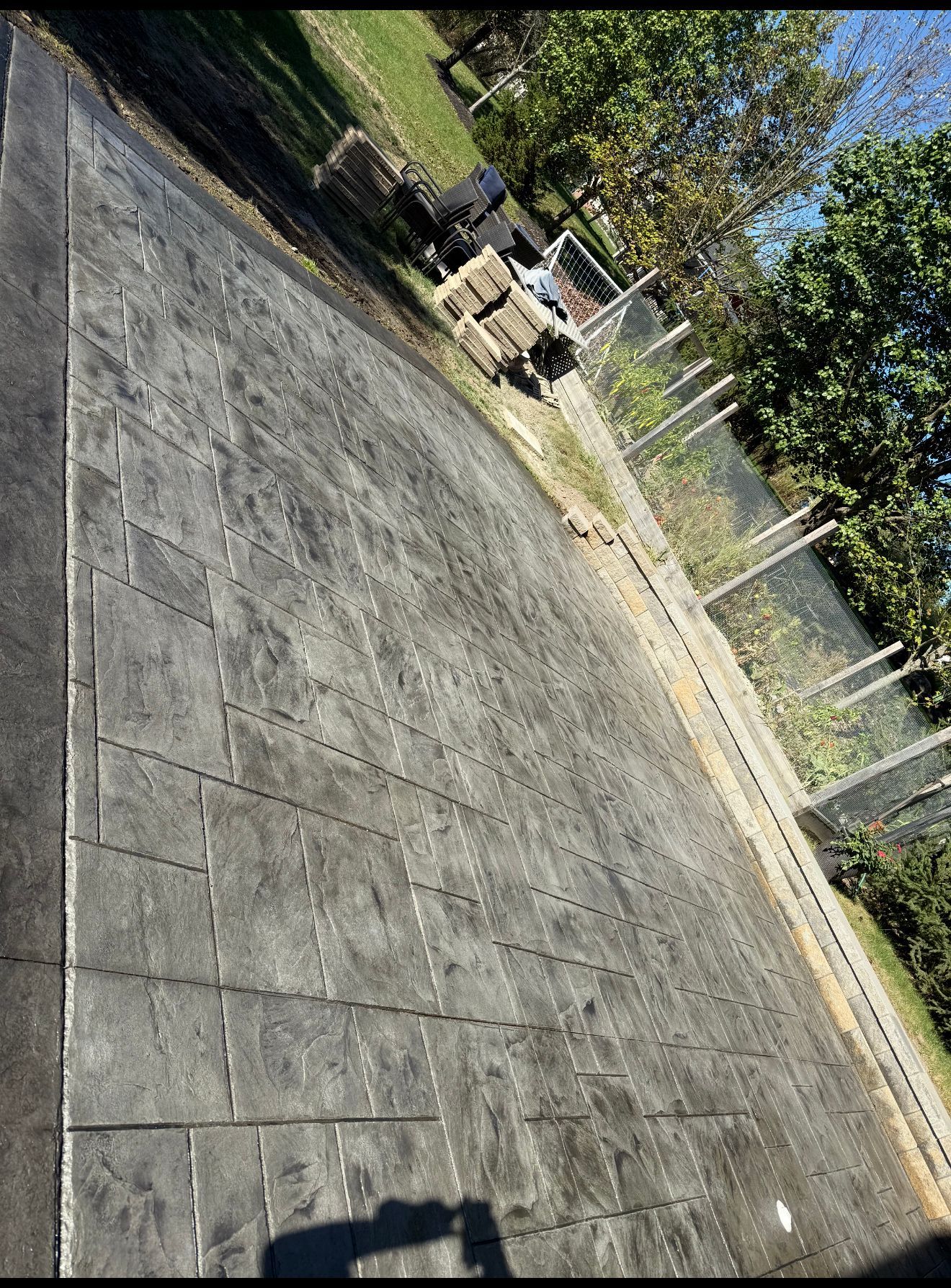 Gray stamped concrete patio with stacked stone and grass, fence, and trees in background.