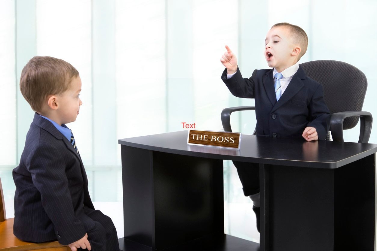 Two boys in suits at a small office desk, one seated and speaking while the other stands nearby.