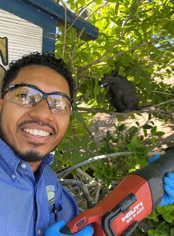 Smiling person in blue shirt holding a red tool, taking a selfie outdoors beside a black cat on a tree branch