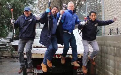 Four people in jackets jump off a stone ledge outdoors, smiling and posing together.