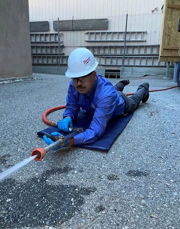 Worker in blue safety gear using a hose to spray water on a gravel surface outdoors.