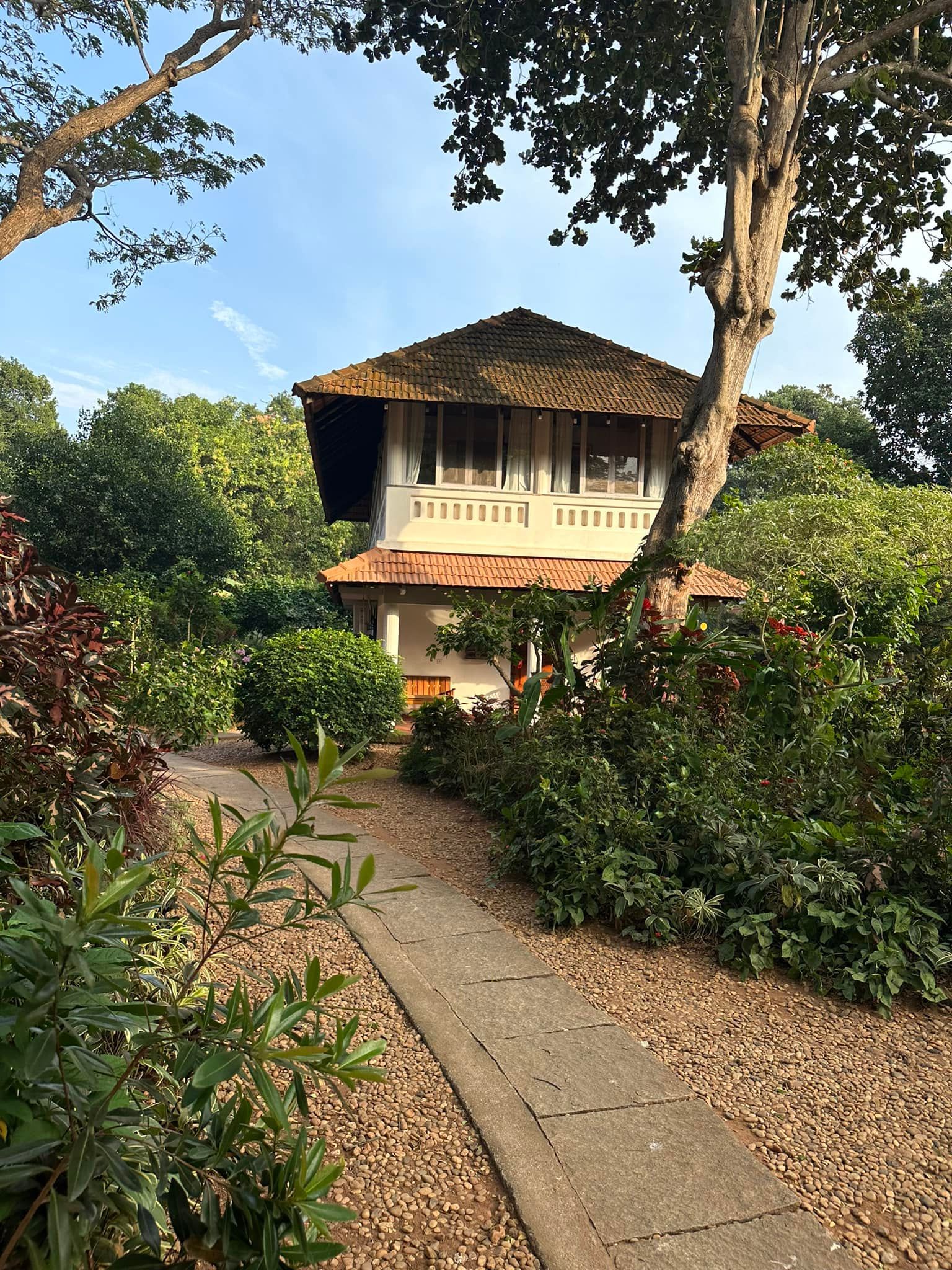 Tropical garden path leading to a two-story house with a tiled roof and balcony under blue sky