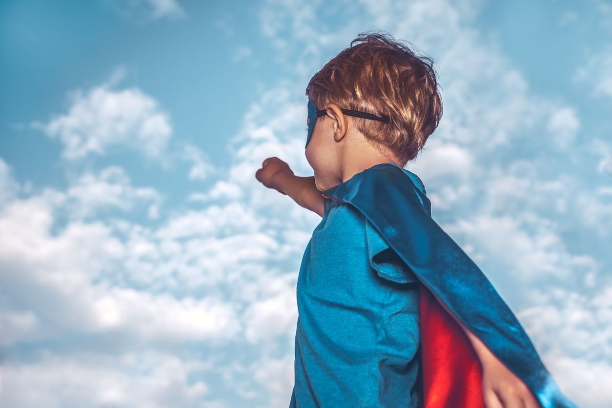Child in a blue shirt and red cape standing against a bright cloudy sky, looking upward.