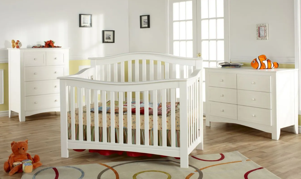 White nursery with a crib, dresser, and chest. Room has a rug and window.