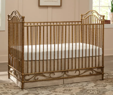 Gold-colored metal baby crib with decorative scrollwork, tan mattress, and matching dust ruffle, in a nursery.