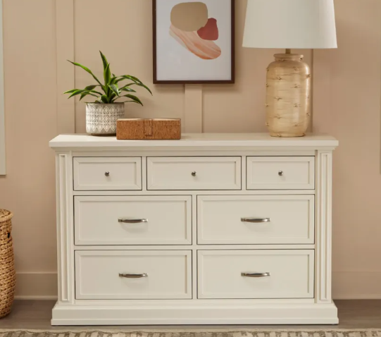White dresser with seven drawers, a lamp, artwork, and a potted plant on top.