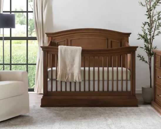 Wooden crib in a room, with a draped blanket and a tall plant.