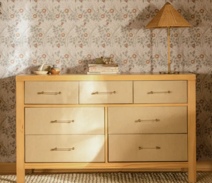 Wooden dresser with seven drawers, lamp, books, and decor against patterned wallpaper.
