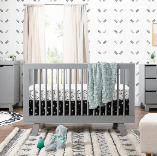 Gray crib in a nursery with matching dresser, patterned rug, and wall decor.