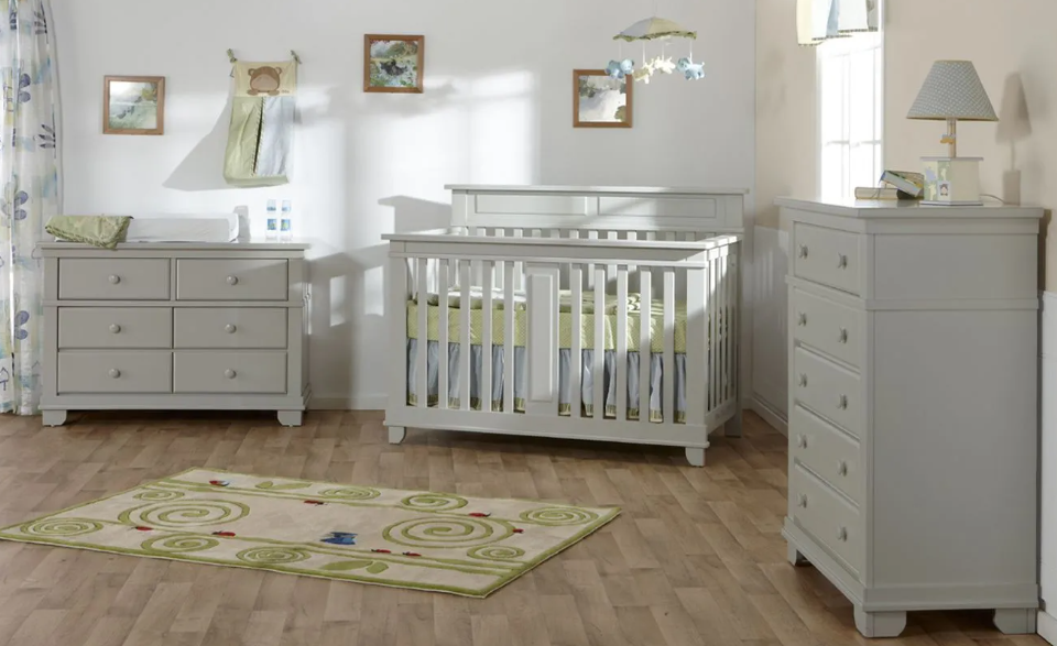 A nursery with a light gray crib, dresser, and chest. Floral rug on wood floor.