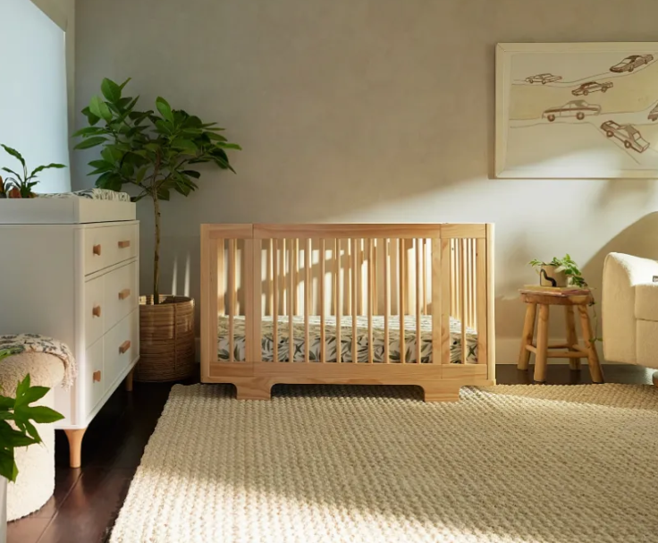 Light wood crib in a neutral-toned nursery with a dresser, rug, and small stool.