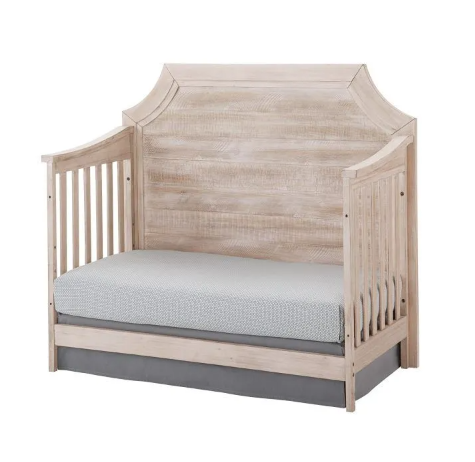Crib converted to a daybed with a light wood finish, white mattress, and gray skirt.