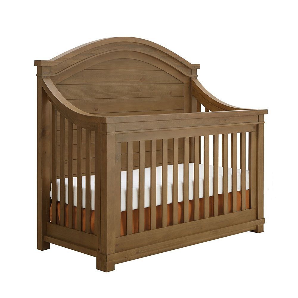 Wooden crib with curved headboard and vertical slats; white mattress and brown skirt.