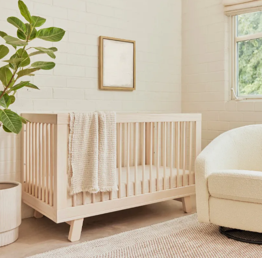 A light-colored crib in a neutral nursery with a blanket, plant, and chair.