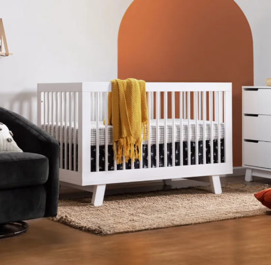 White crib in a nursery, with a yellow blanket, and a white dresser.