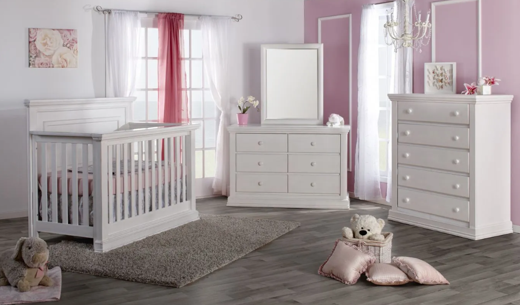 White nursery furniture set in a pink room: crib, dresser, mirror, chest of drawers, rug, and decorative items.