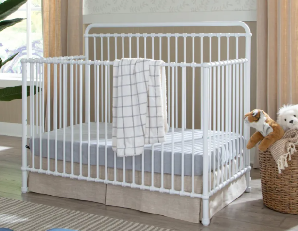 White metal crib with a mattress and blanket; next to a wicker basket with stuffed animals.